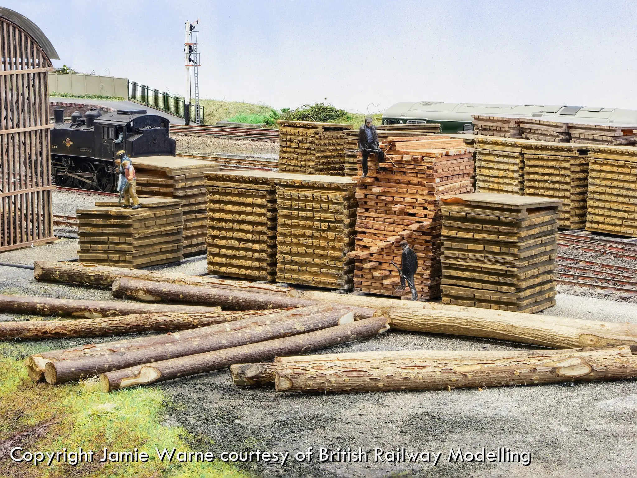 Stacks of sleepers and logs in the Redbridge works yard, with a steam locomotive shunting behind